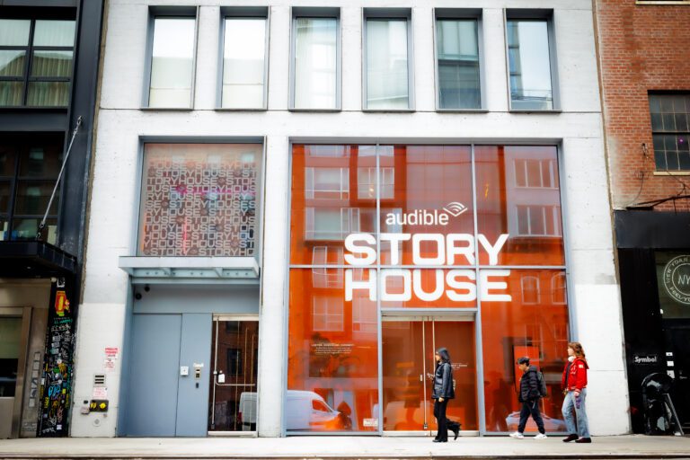 Urban storefront with bright orange glass displaying 'AUDIBLE STORY HOUSE'; pedestrians walk by on the sidewalk.