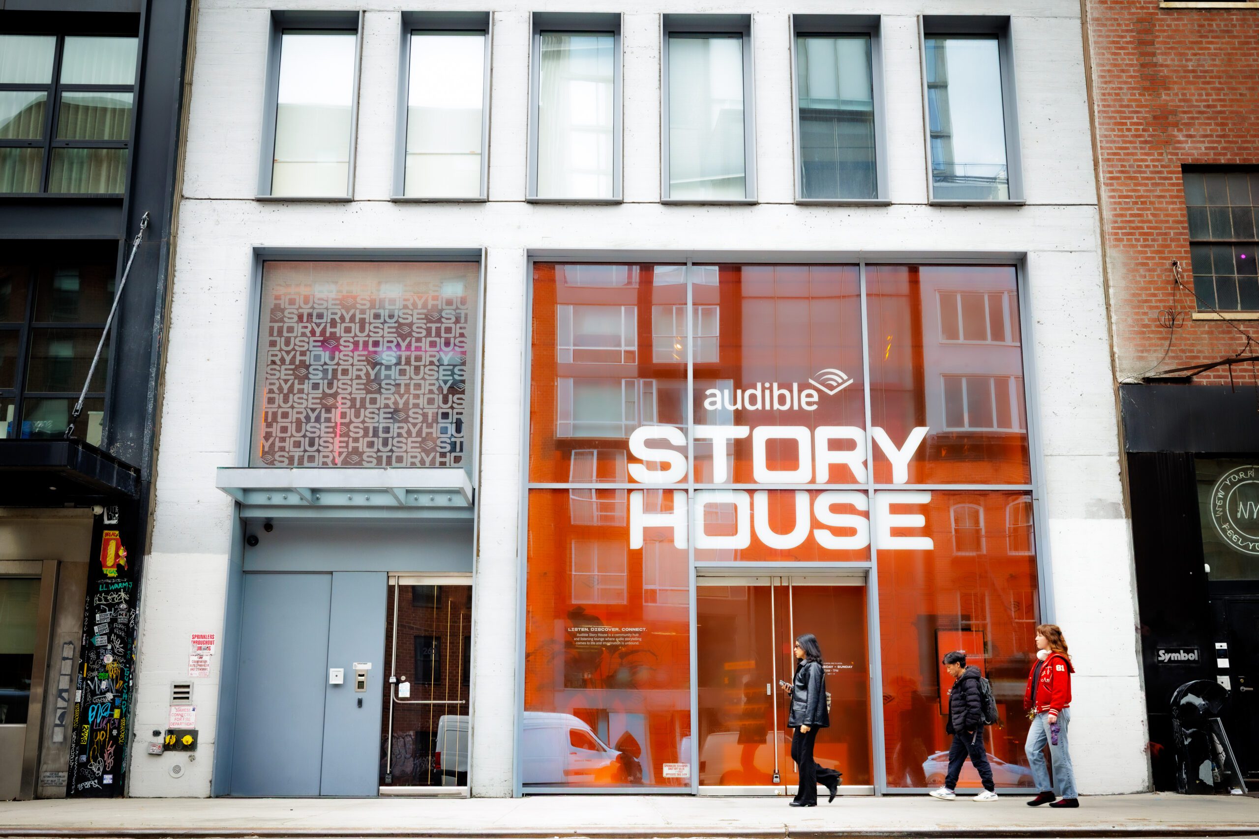 Urban storefront with bright orange glass displaying 'AUDIBLE STORY HOUSE'; pedestrians walk by on the sidewalk.