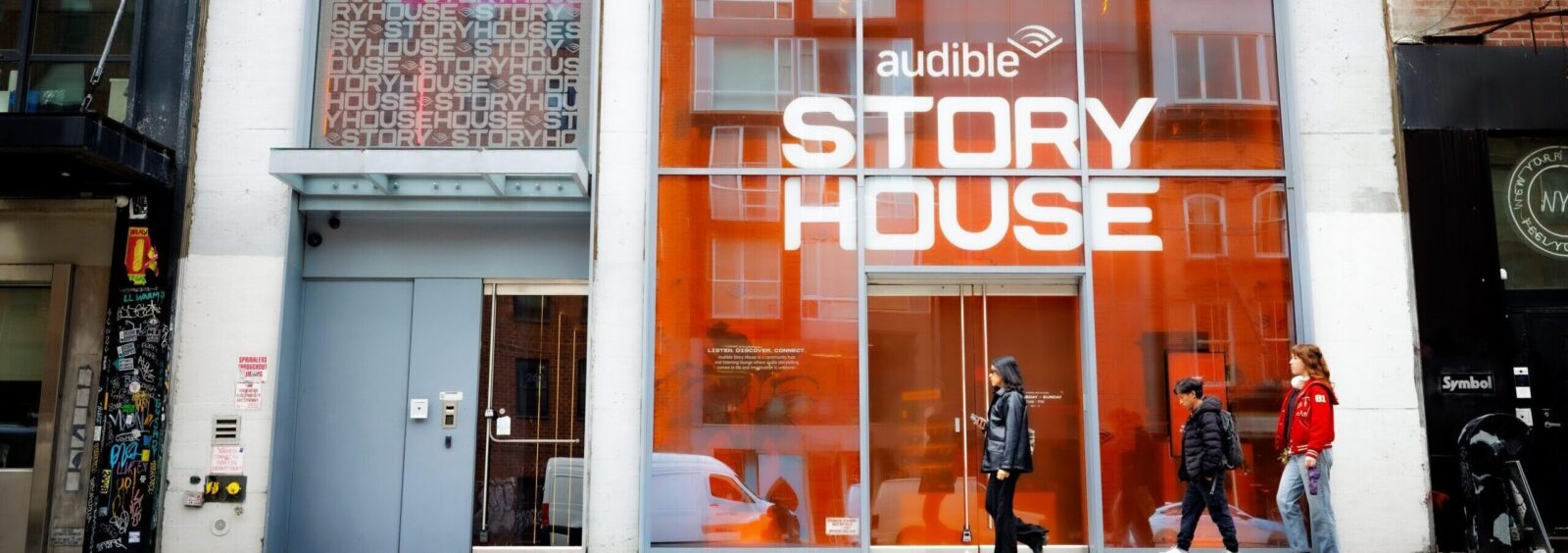 Storefront with orange-tinted glass, Audible logo, and large 'STORY HOUSE' lettering; pedestrians walking by on a city sidewalk.