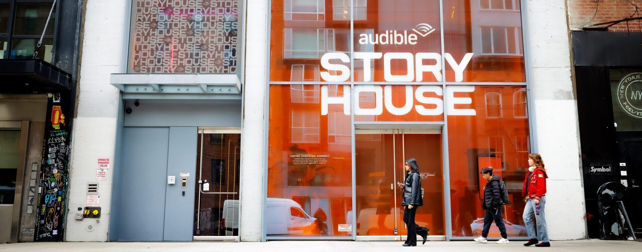 Storefront with orange-tinted glass, Audible logo, and large 'STORY HOUSE' lettering; pedestrians walking by on a city sidewalk.