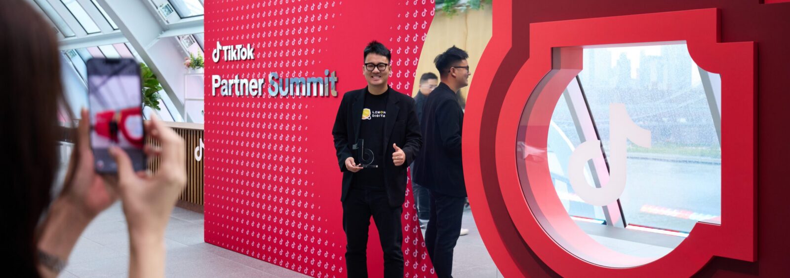 Man posing for a photo in front of a red TikTok Partner Summit backdrop; a person in the foreground holds a smartphone to take the picture.