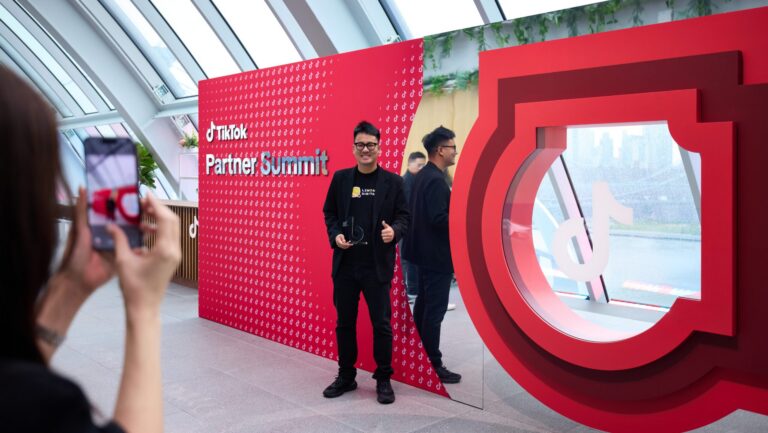 Man posing for a photo in front of a red TikTok Partner Summit backdrop; a person in the foreground holds a smartphone to take the picture.