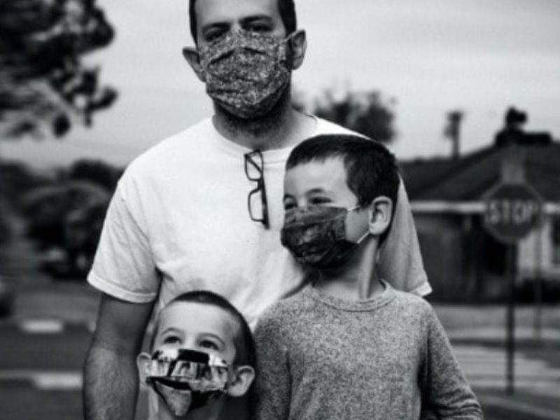Man and his sons stand together, masks on, in a somber black and white toned photo